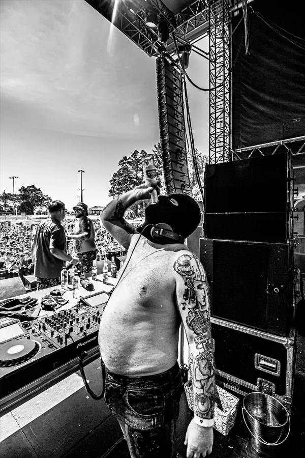 A tattooed, shirtless Bad Boy Chiller Crew member on stage at an outdoor music festival, pouring a drink, with the festival crowd in the background.
