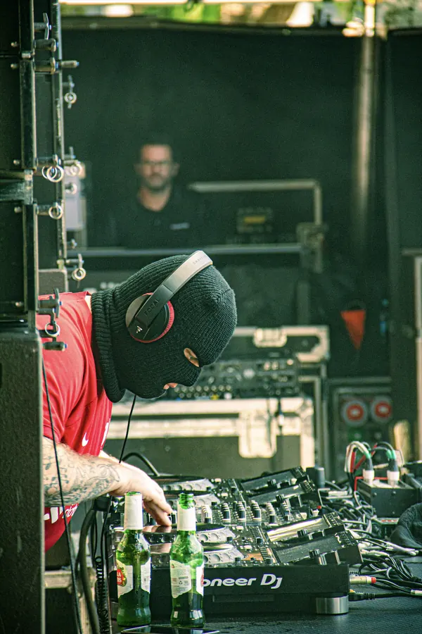 A Bad Boy Chiller Crew DJ wearing a black ski mask, red shirt and headphones, working at a DJ booth with a mixer and bottles of beer.