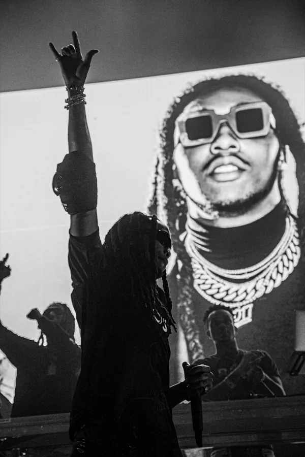 A performer with dreadlocks and sunglasses holding a microphone on stage in front of a large screen showing a close-up of a man wearing sunglasses and chains.