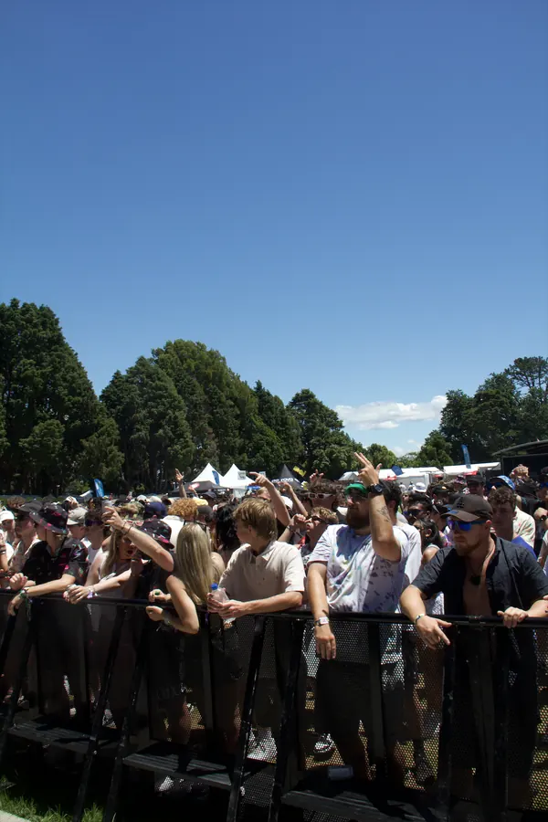 Crowd at Bay Dreams standing behind the black metal barrier, some raising their arms or holding water bottles, under a clear blue sky surrounded by trees.