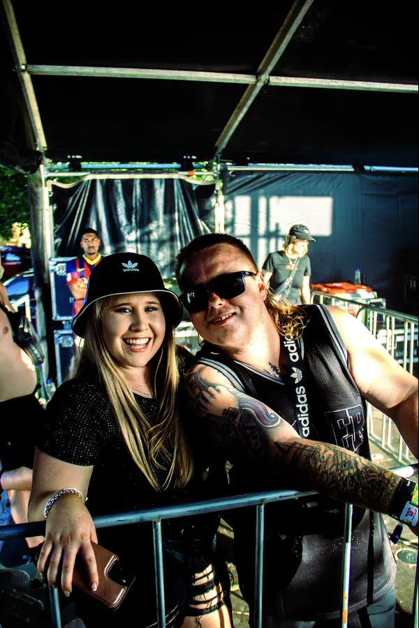 A woman and a man smiling behind the barrier at a music event. She wears a black Adidas hat and holds a smartphone; he has tattoos, sunglasses and a black sleeveless Adidas shirt.