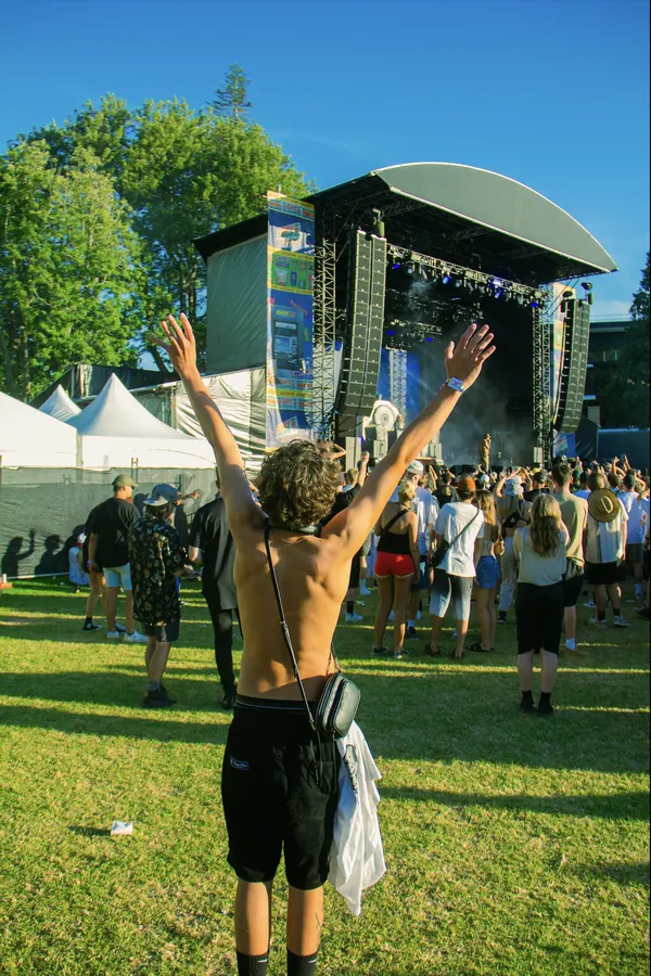 A shirtless festival-goer with curly hair raising their hands in front of the crowd at Bay Dreams, with the main stage and blue sky behind.