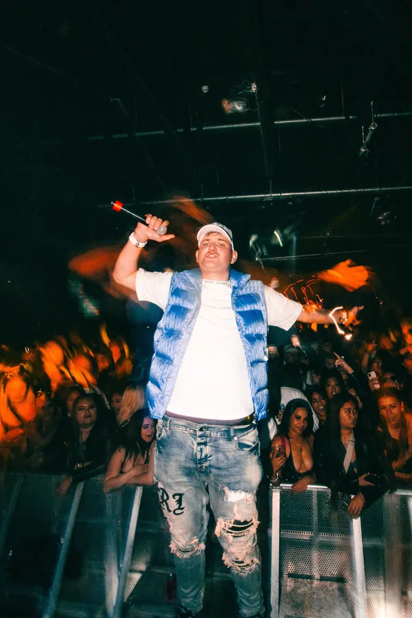 Performer in a blue puffer vest holding the mic up to a packed club crowd.