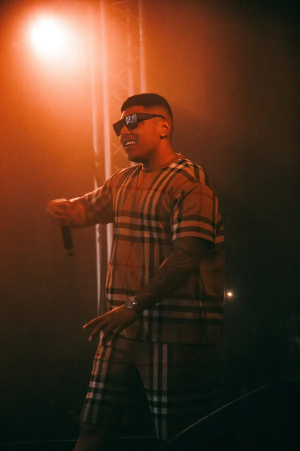 Performer in a plaid shirt and sunglasses under orange stage light.