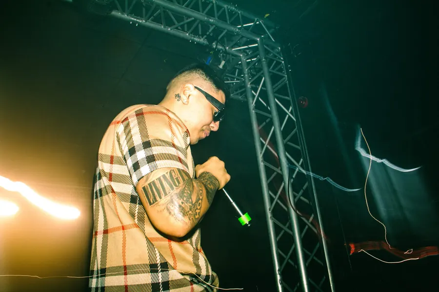 Performer in a plaid shirt rapping on stage beneath the lighting truss.