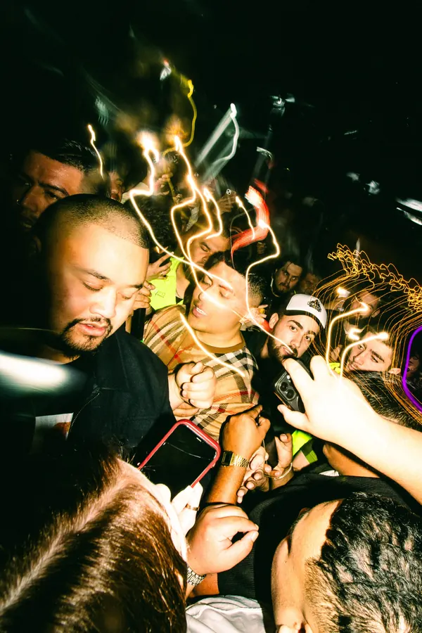 A rapper surrounded by fans in a packed club, with phone flashes and light trails around him.
