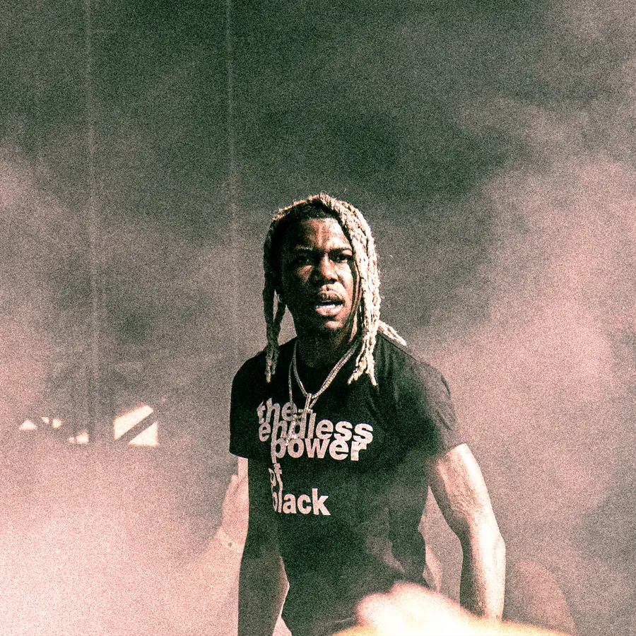 Destroy Lonely with blonde dreadlocks, wearing a black t-shirt with white text, standing in a smoky environment with a serious expression, looking into the camera.