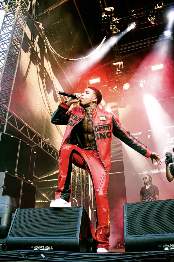 NLE Choppa rapping into a microphone on stage at Bay Dreams 2024, wearing a red and black leather jacket and matching red pants, with stage lights, speakers and DJs around him.