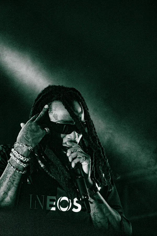 Quavo with dreadlocks and sunglasses performing on stage, holding a microphone in one hand and touching his head with the other, wearing rings and bracelets.