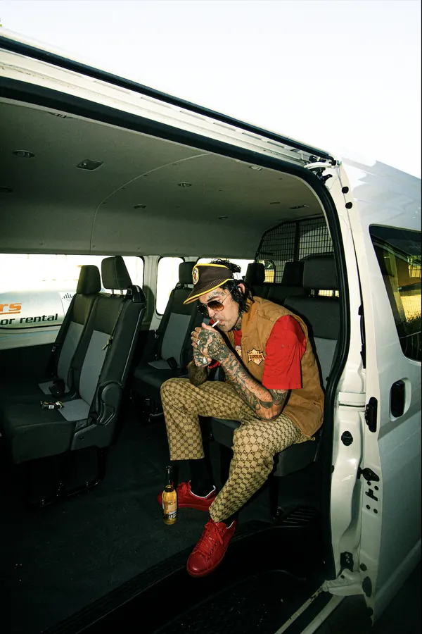 Yelawolf sitting inside a white van, wearing sunglasses, a brown vest over a red shirt, patterned pants and red shoes, with a beer bottle on the floor.